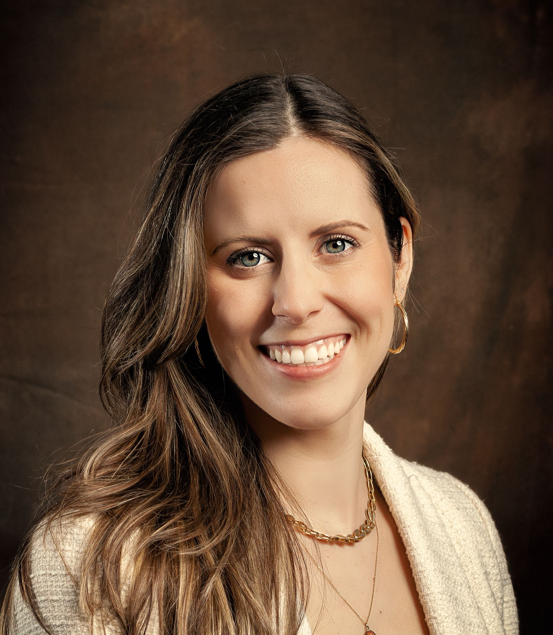 Headshot of a person with long wavy hair, smiling, wearing a cream textured blazer and layered gold necklaces.
