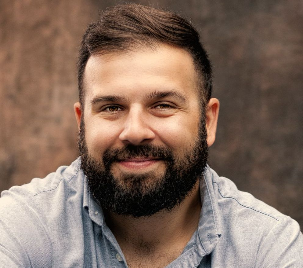 A person with a beard and short dark hair smiling, wearing a light blue button-up shirt against a textured brown backdrop.