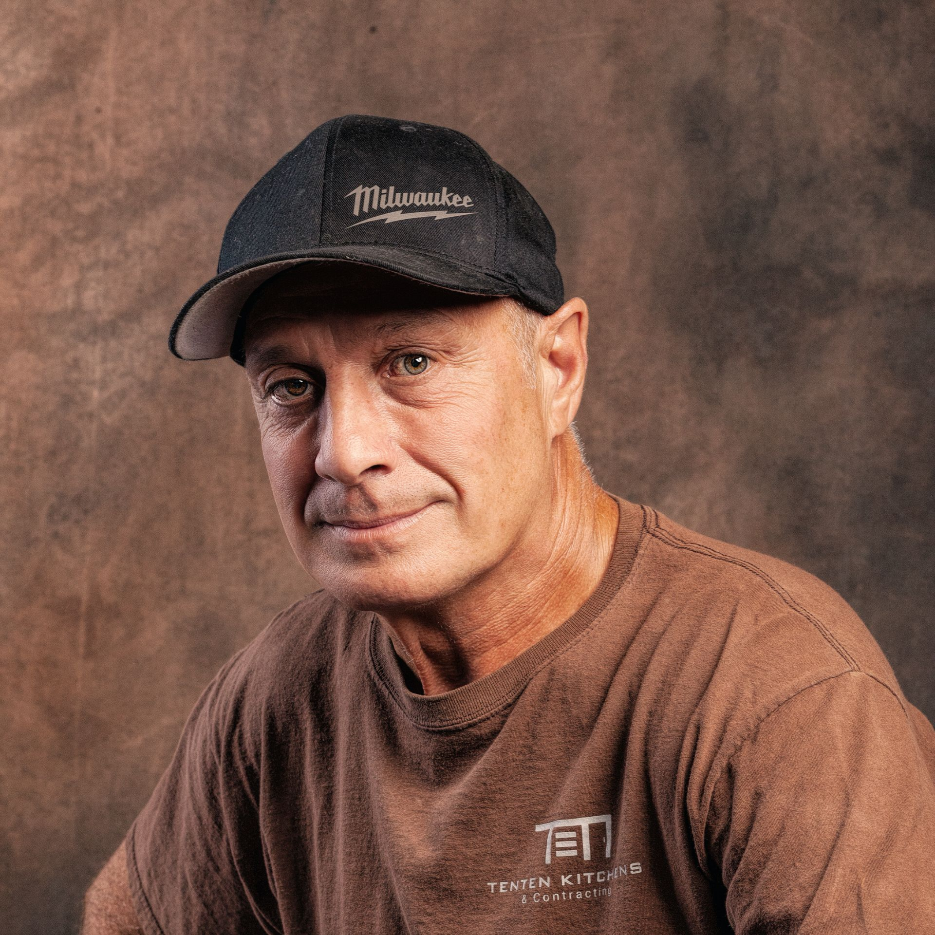 A person wearing a dark baseball cap and a brown t-shirt with a logo, posing against a dark, mottled background.