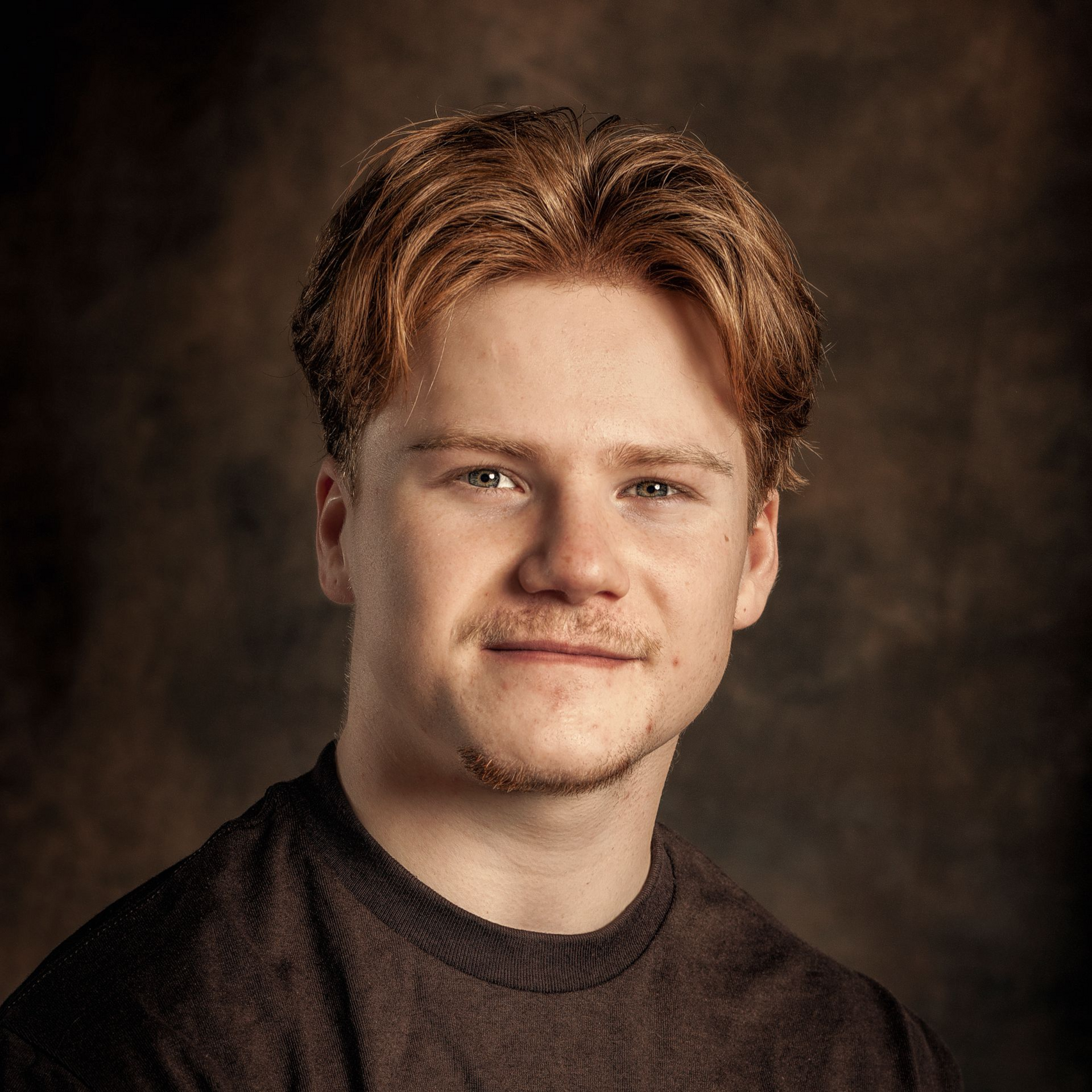 Portrait of a person in a dark shirt against a brown studio backdrop, looking at the camera.