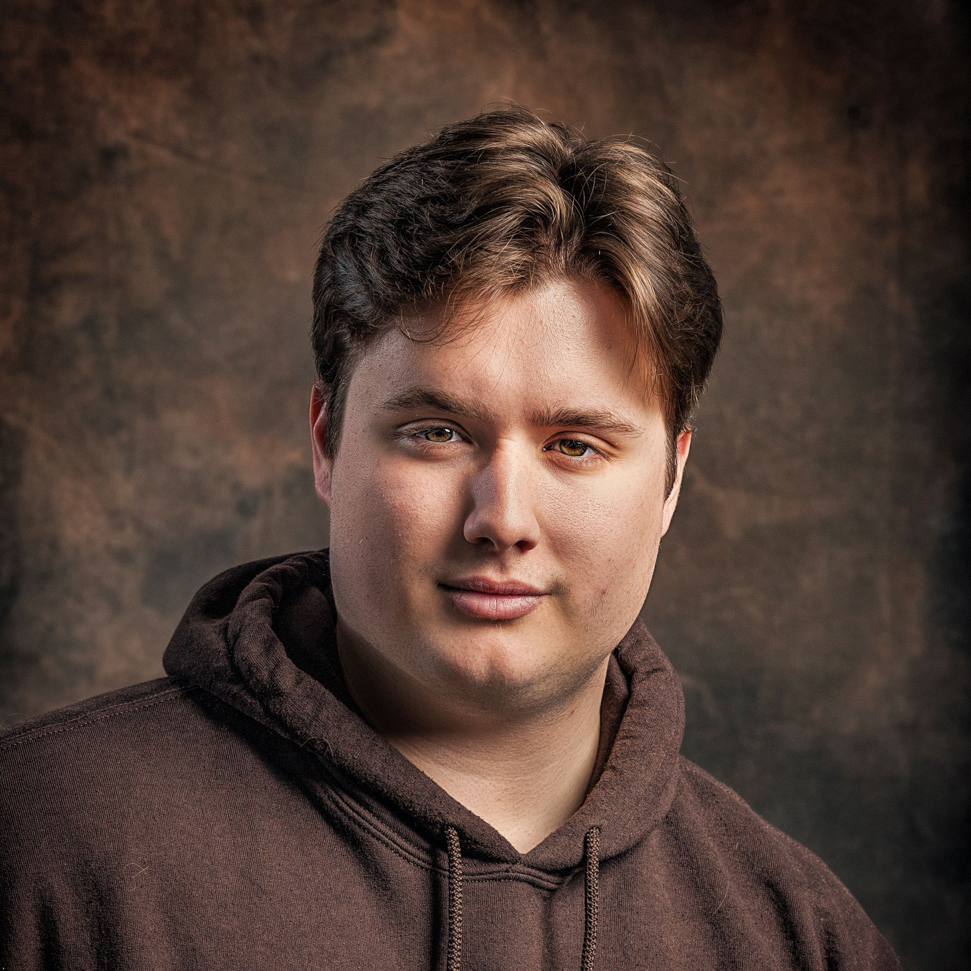 A person with short brown hair wears a brown hooded sweatshirt against a textured, dark brown mottled background.