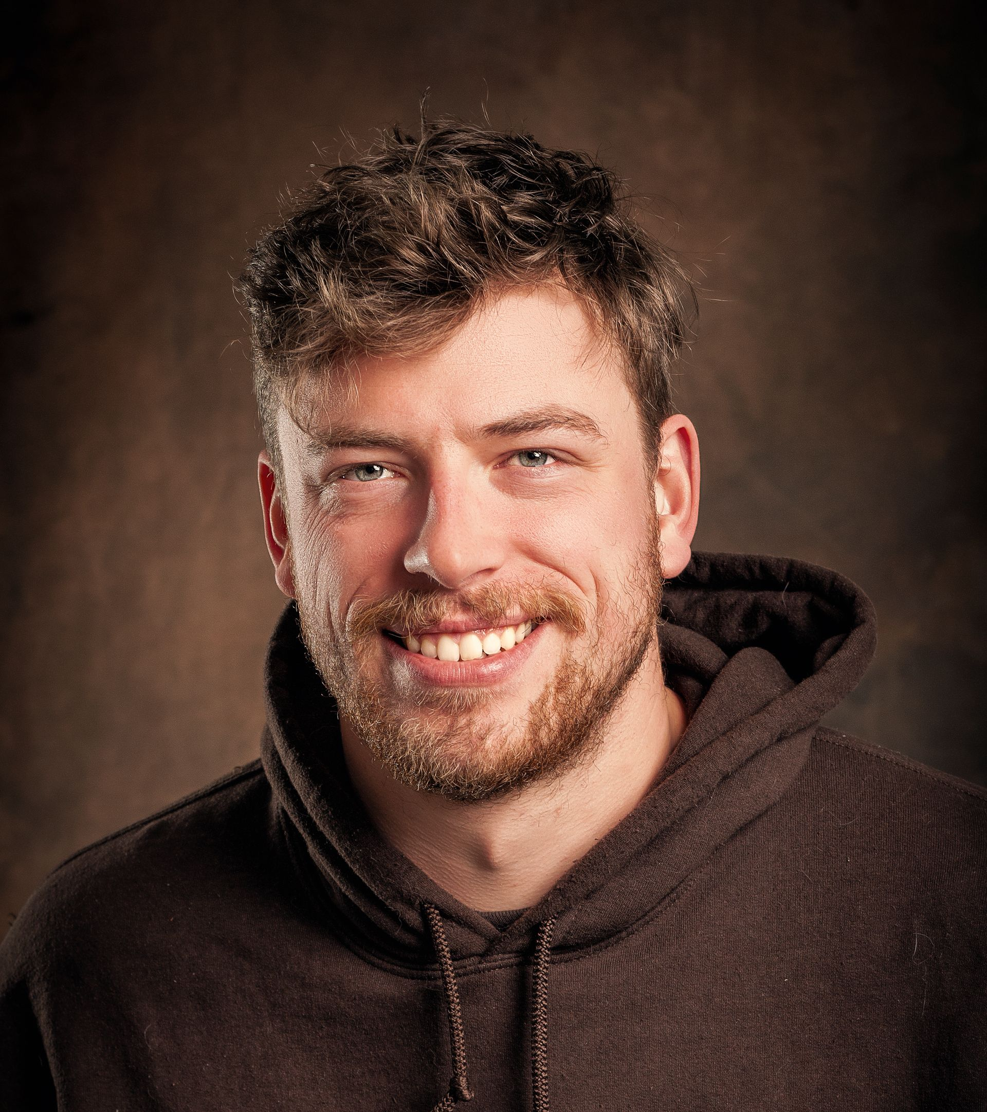 A person with short, wavy hair and a beard, smiling, wearing a dark brown hoodie against a dark, textured background.