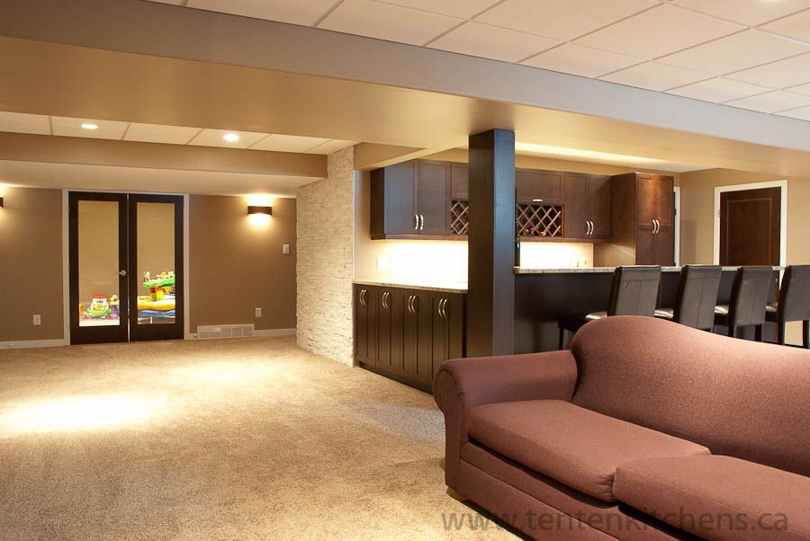 A finished basement with a brown sofa in the foreground, a home bar area with stools, and double glass doors.