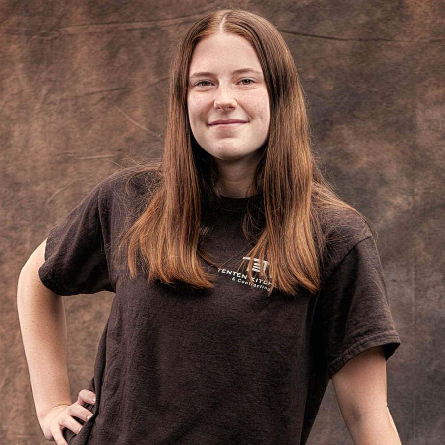 A smiling person with long brown hair wearing a brown t-shirt and a necklace, posed against a dark, textured background.
