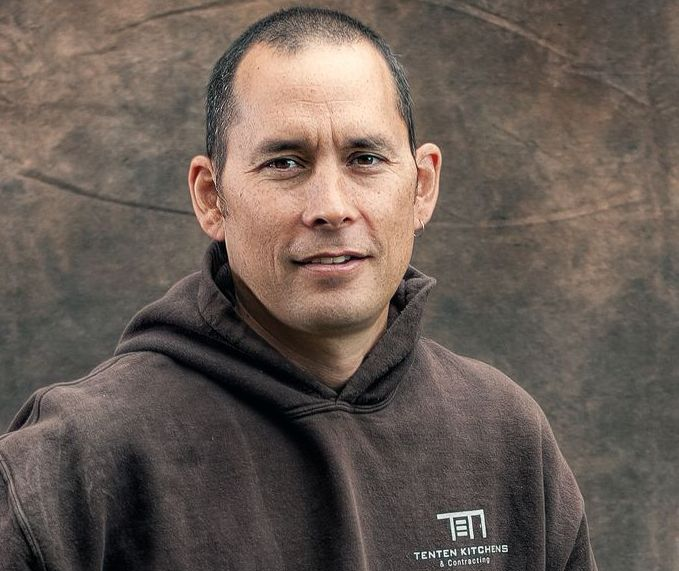 A person with a neutral expression wearing a brown hoodie with a white logo, set against a dark, textured background.