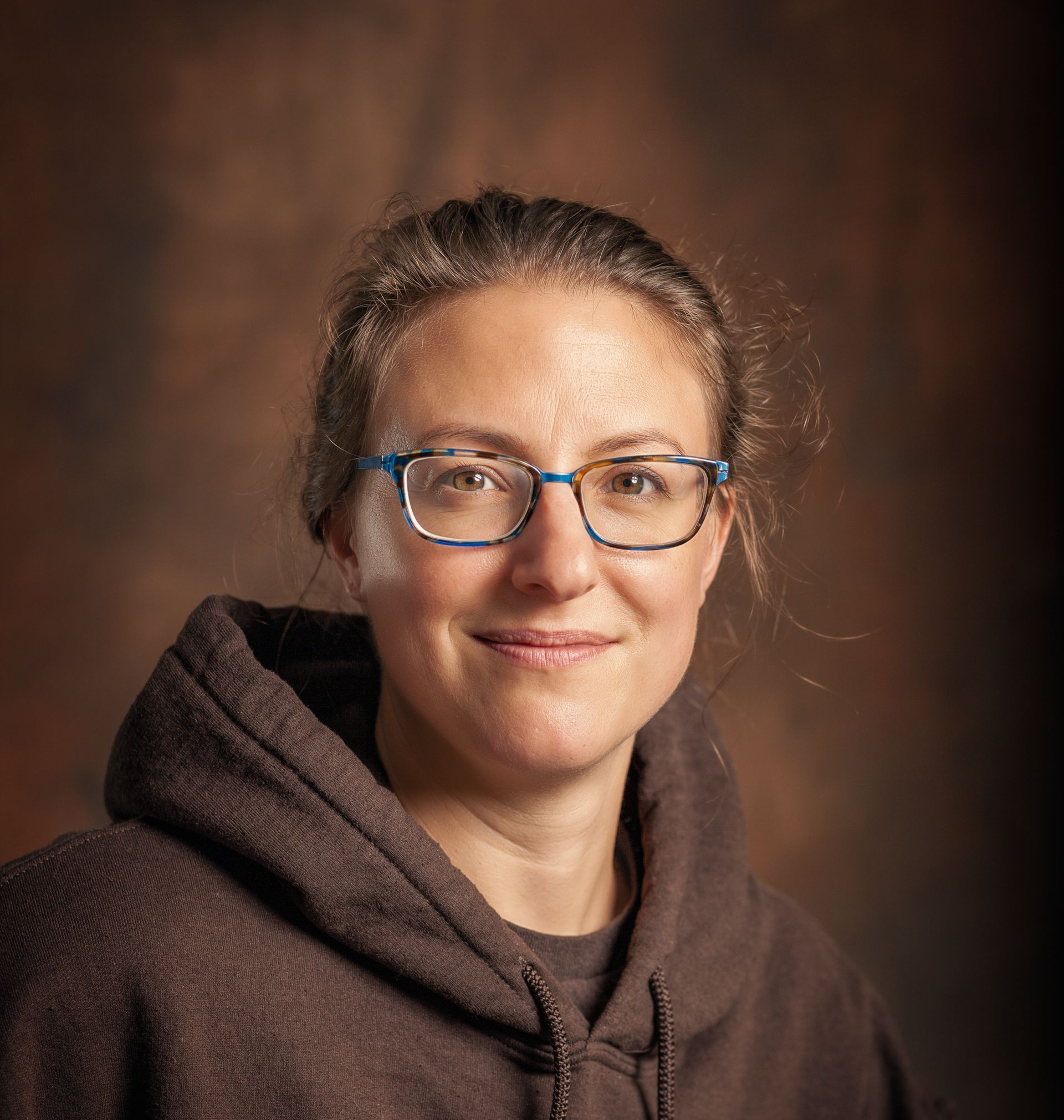 A person with glasses wears a brown hooded sweatshirt in a headshot against a textured brown background.