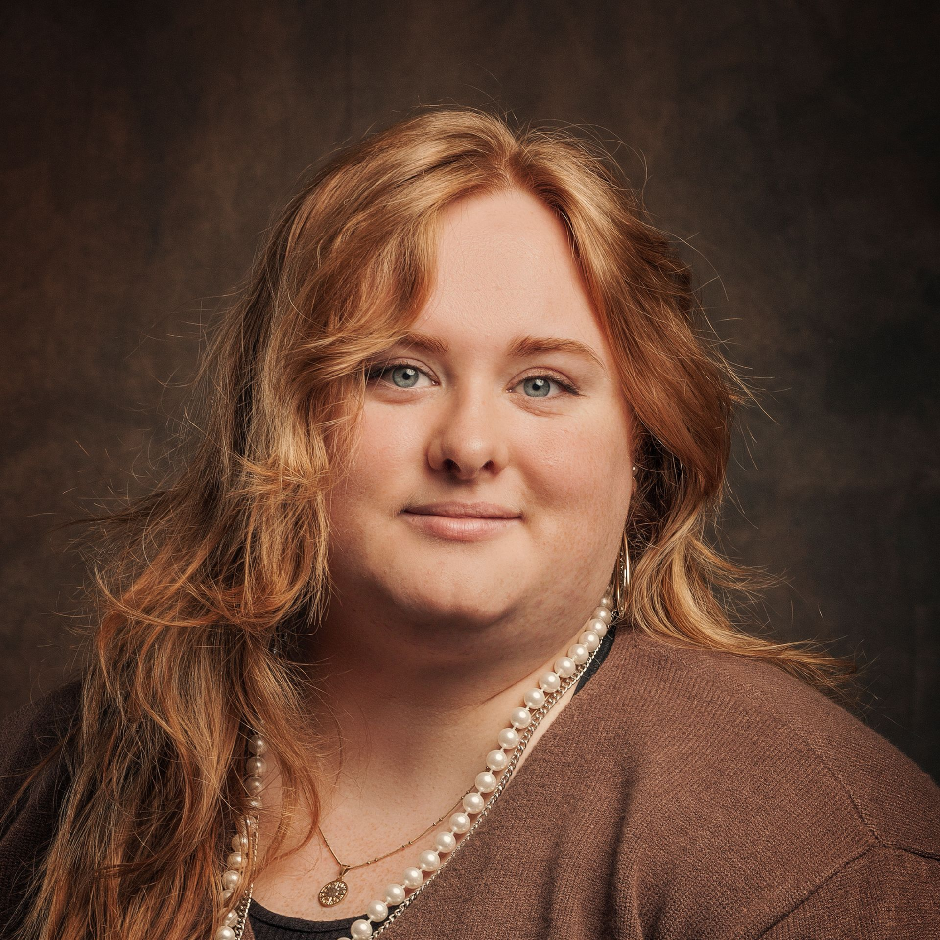 Portrait of a person with long hair wearing a brown top and layered necklaces against a dark background