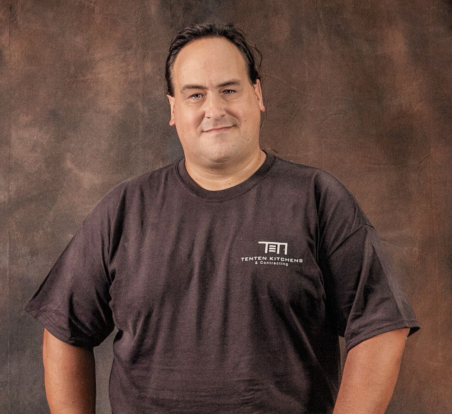 A man wearing a dark brown t-shirt with a white graphic stands against a mottled, textured brown background.