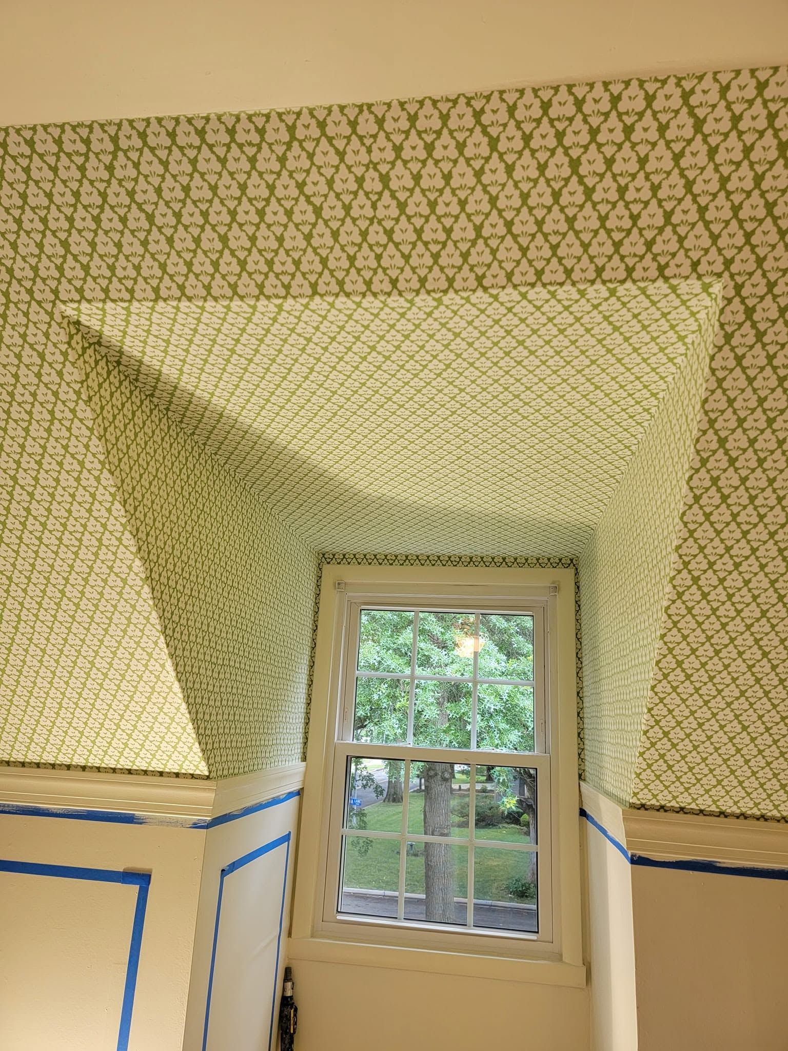 Small window in a room with yellow patterned walls and blue painter’s tape; trees visible outside.