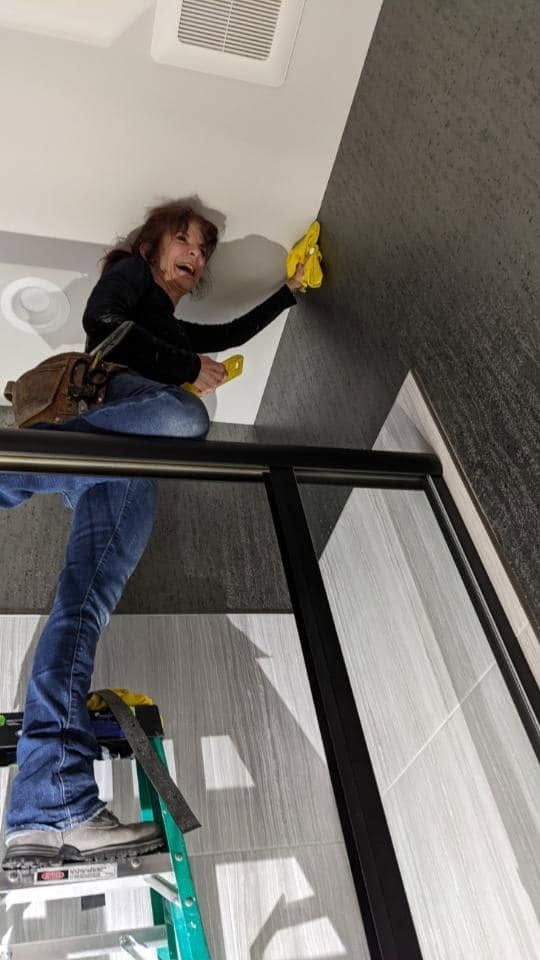 Person in black top and jeans cleaning a textured wall with a yellow cloth, standing on a ladder.