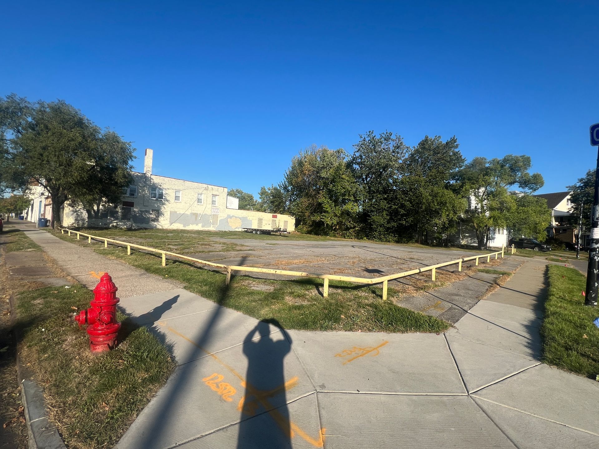 Empty lot enclosed by yellow railing at a street corner, red fire hydrant, bright blue sky.