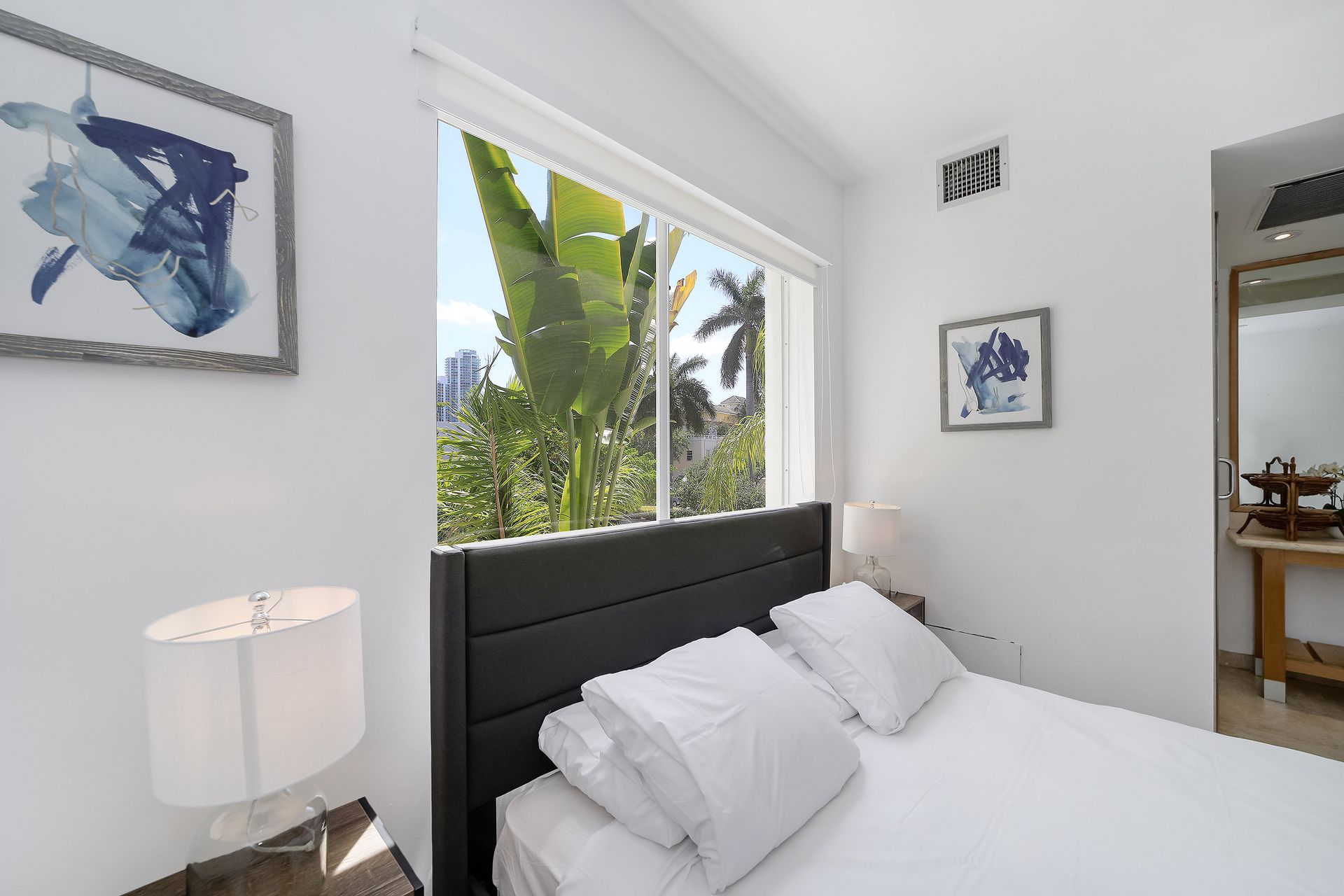 Bedroom with a bed, artwork, a window overlooking greenery, and a table with a mirror.