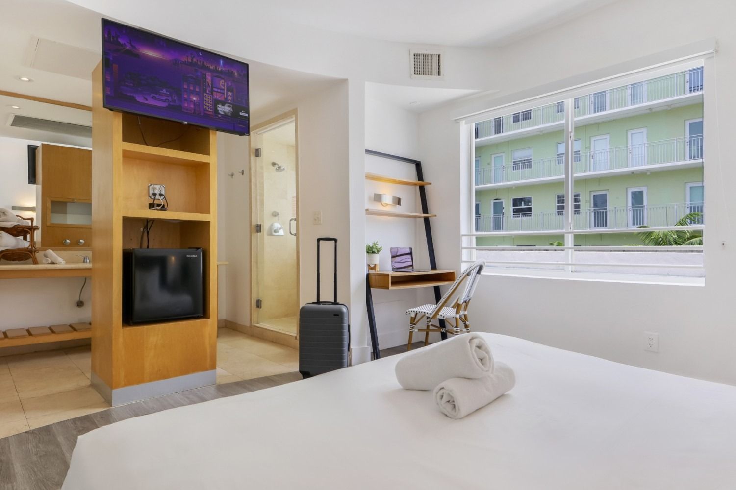 Hotel room with bed, TV, luggage, desk, and view of a green building.