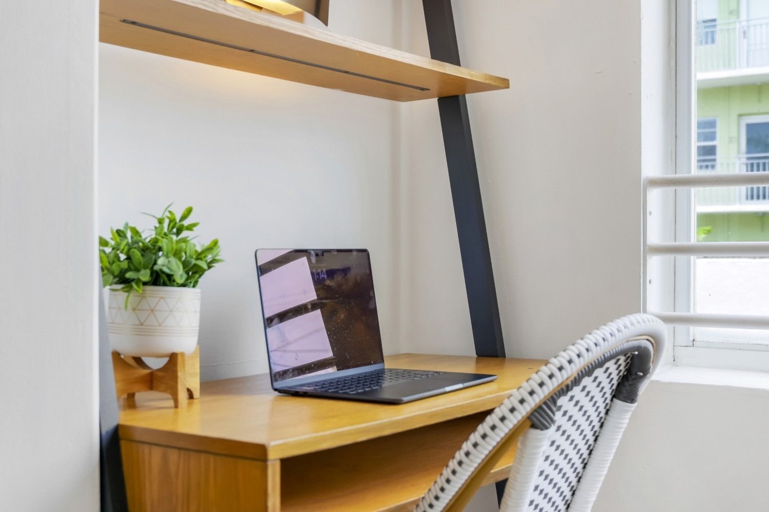 Desk with laptop, plant, and chair by a window. Wooden desk and shelf against a white wall.