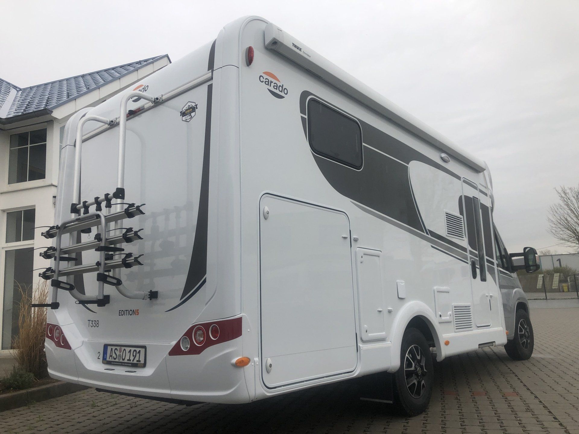 Ein weißes Wohnmobil mit einem Fahrradträger auf der Rückseite ist vor einem Gebäude geparkt.