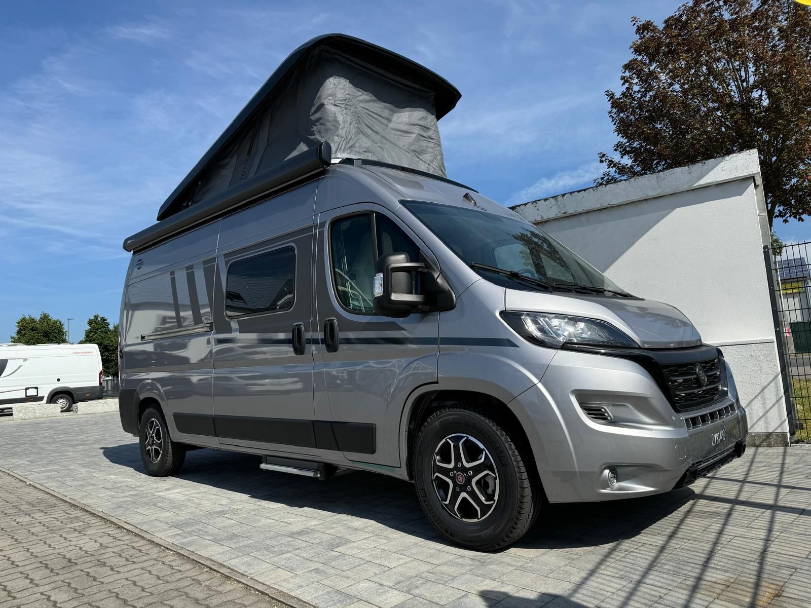 Grauer Campervan mit Aufstelldach parkt an einem sonnigen Tag draußen.