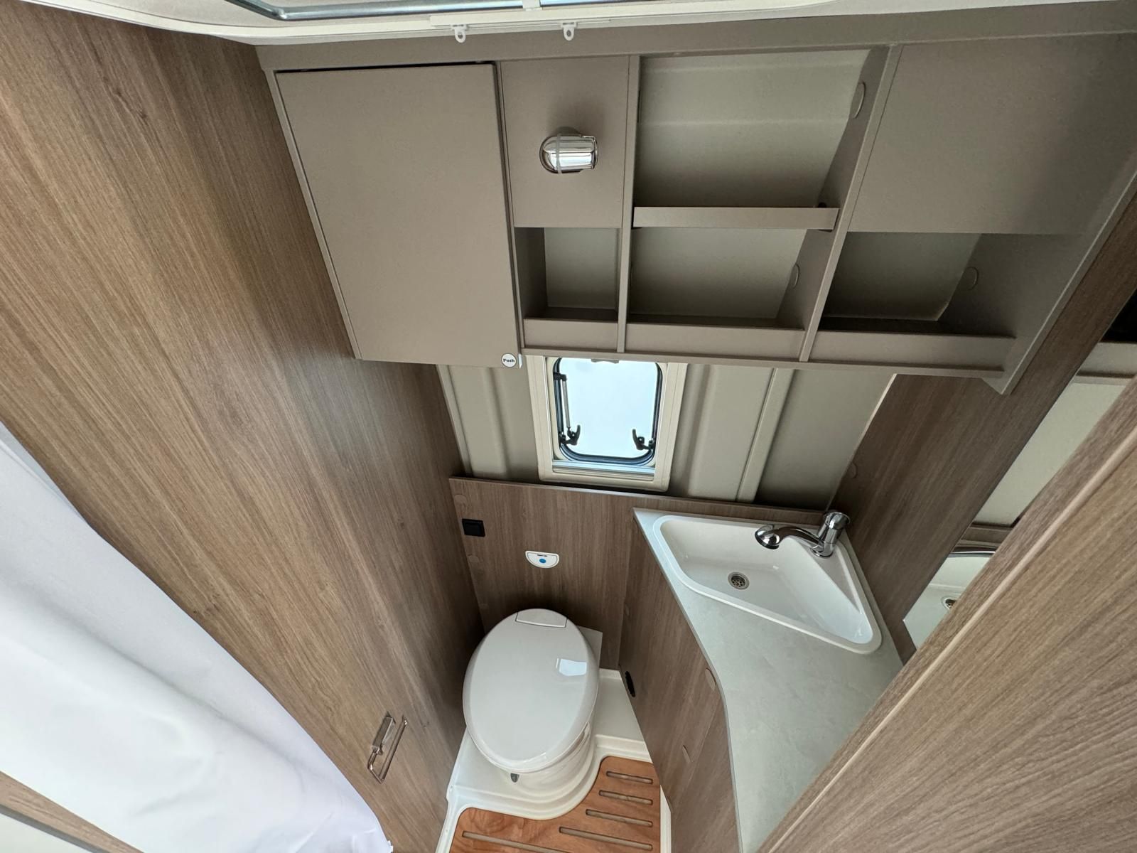 Innenansicht eines Wohnmobil-Badezimmers: Toilette, Waschbecken, kleines Fenster, Regale und hellbraune Holzvertäfelung.