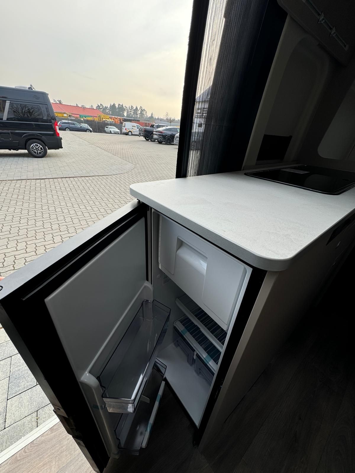 Kühlschrank im Inneren eines Lieferwagens mit offener Tür. Helle Arbeitsfläche und Blick auf geparkte Autos draußen.
