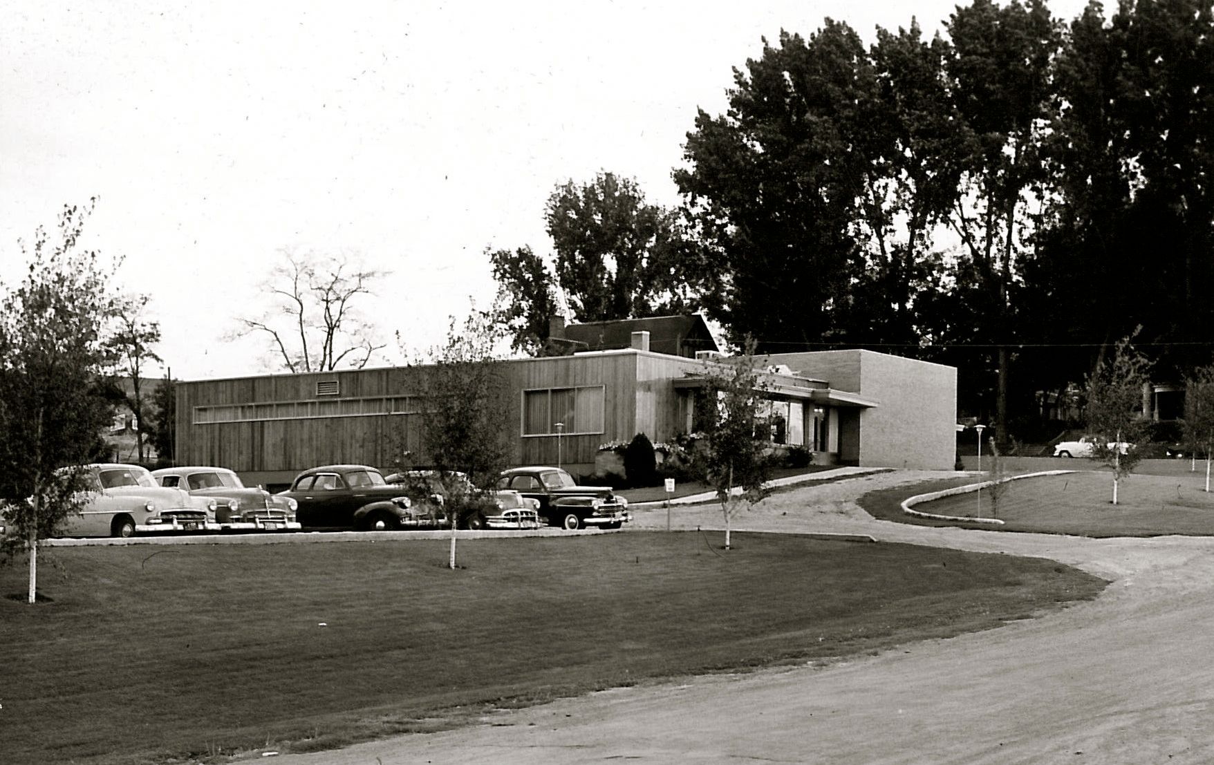 Black-and-white photo of a low modern building with parked cars and a curved driveway under trees.