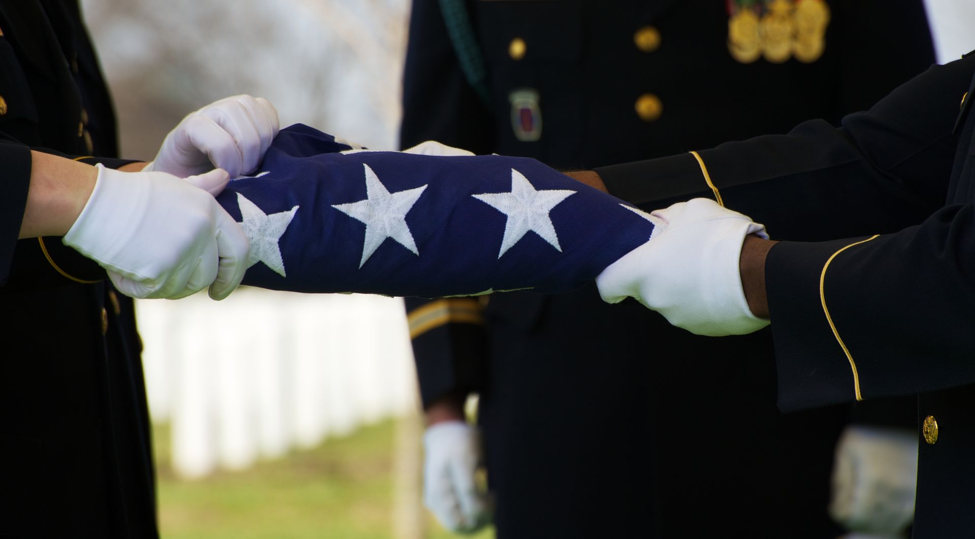Hand resting on a flag-draped coffin beside white flowers at a graveside funeral