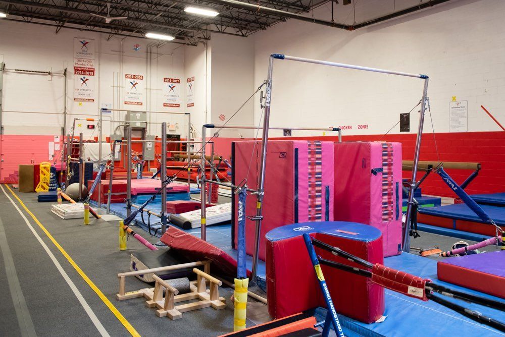 Gymnastics training facility with various apparatus, including bars, beams, and mats in pink, red, and blue.