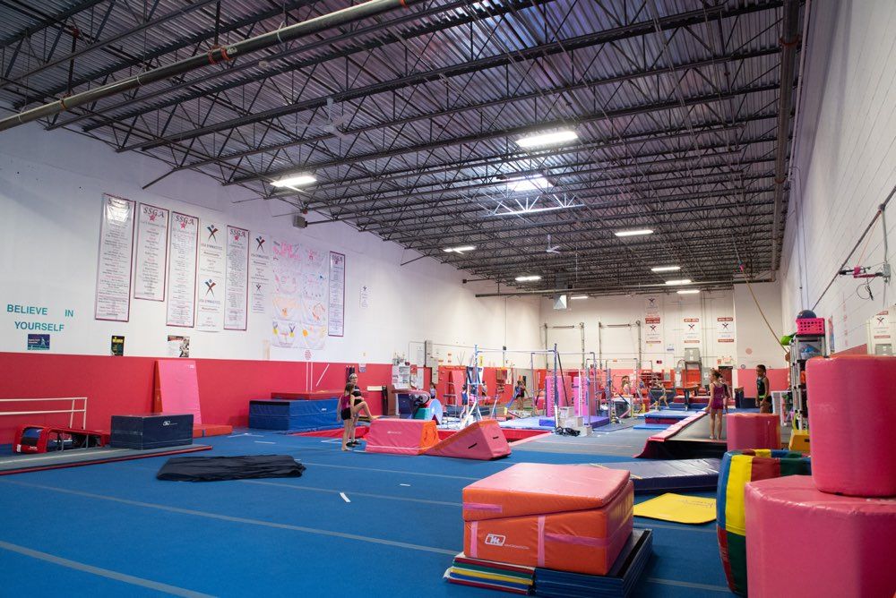 Gymnasium interior with gymnastics equipment on blue mats, red and white walls, and a high ceiling.