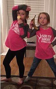 Two girls in pink shirts pose; one eats a banana, the other holds one to her ear. Indoors.