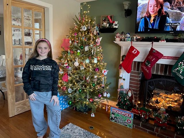 Young person in front of a Christmas tree and fireplace, inside a home. The person is smiling.