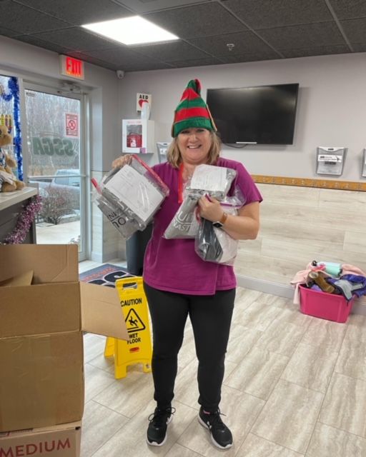 Woman in elf hat holding bags of donations in an office setting. She's smiling, near a box and doorway.