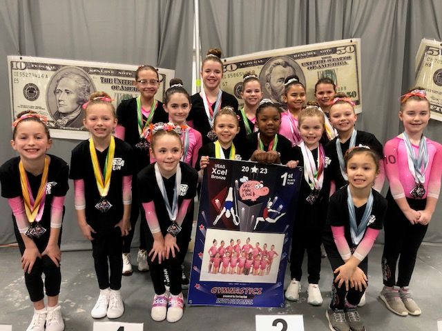 Gymnastics team poses with medals and a trophy in front of a backdrop with a large dollar bill graphic.
