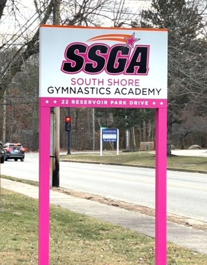 Sign for South Shore Gymnastics Academy, pink and white with logo, on a street.