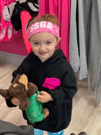 Young child in black hoodie and pink headband holding a stuffed animal. Pink shop backdrop.