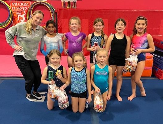 A group of young gymnasts and a coach pose in a gym, holding gifts and smiling.
