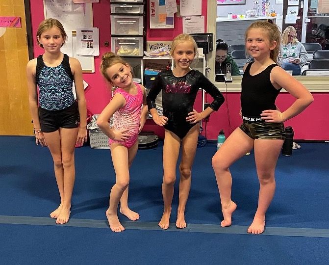 Four young gymnasts in leotards posing on a blue mat in a gym.