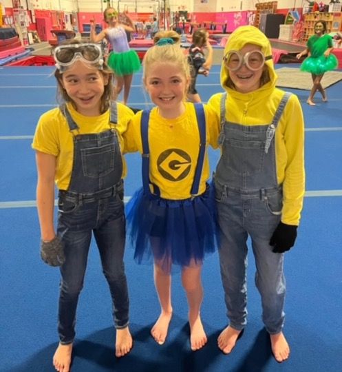 Three children in yellow Minion costumes, one with a blue tutu, in a gym.