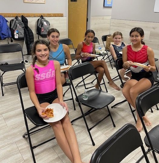Gymnasts in colorful leotards eat pizza on chairs in a room, smiling.