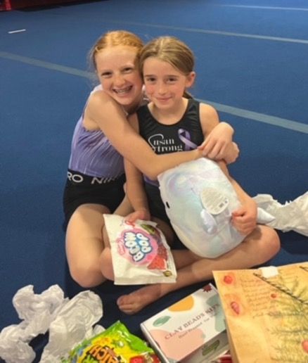 Two girls sit on a blue mat, hugging and smiling, with gifts and a plush toy.