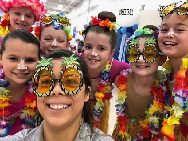 Group of children and an adult wearing leis and pineapple sunglasses, smiling in a gymnasium.