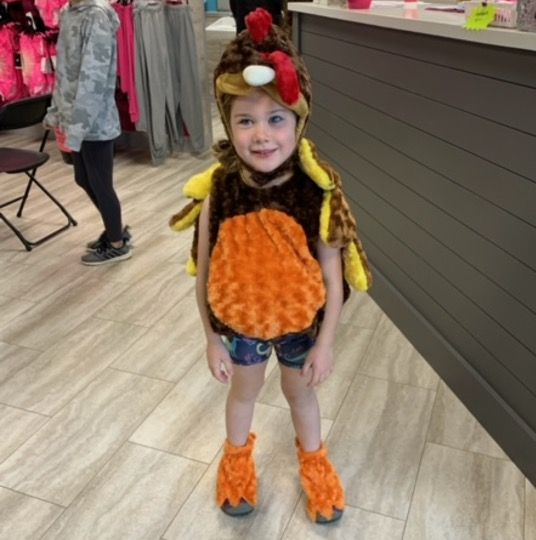 Child in a turkey costume smiles, inside a store. They wear a brown and orange plush outfit.