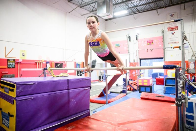 Gymnast on uneven bars in a gym; colorful leotard, legs extended, holding position.