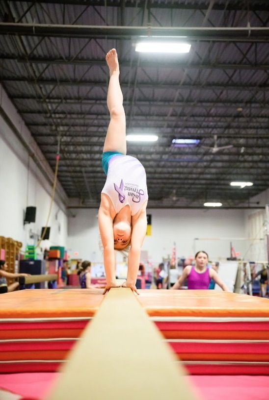Gymnast balances on a beam, in a handstand with one leg extended. The setting is a gymnasium.