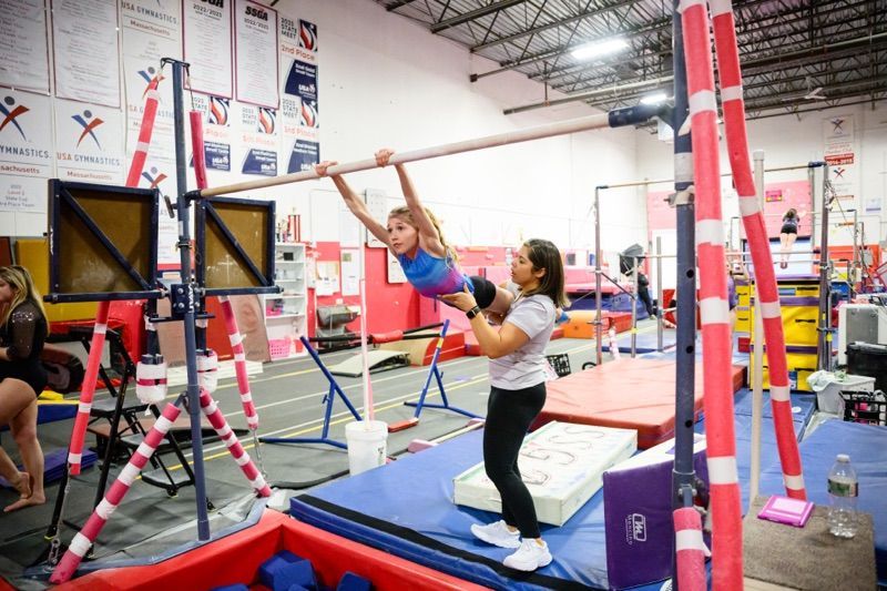 Gymnast on uneven bars, assisted by a coach. Gym interior, blue mats, red and pink equipment.