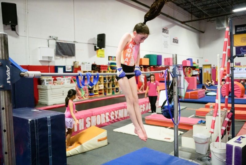 Girl doing a gymnastic bar exercise in a gym. She is hanging with arms straight.