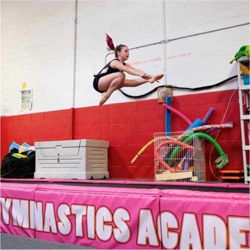 Gymnast in black leotard leaps over obstacles in a pink and red gymnastics academy setting.