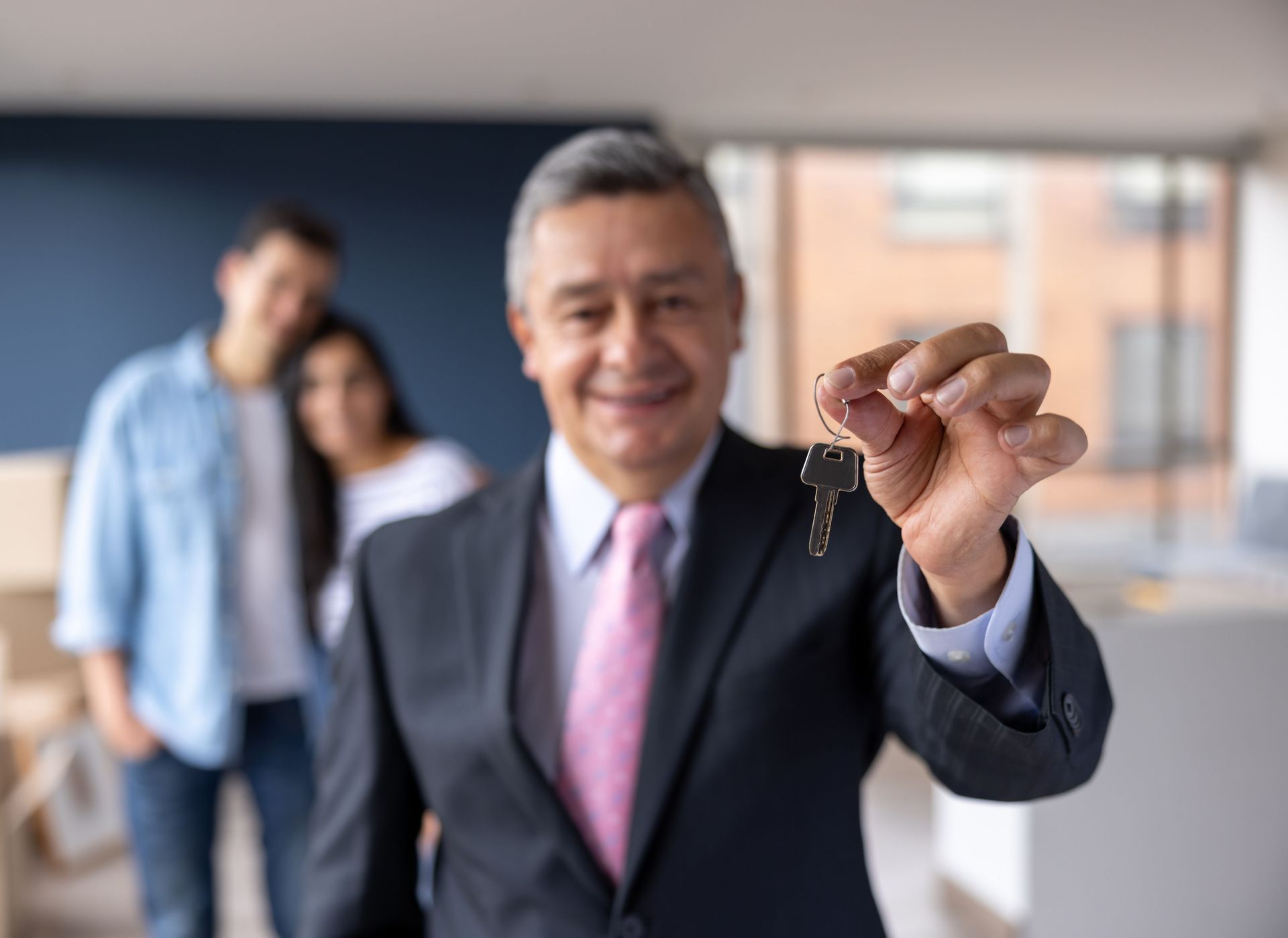 Real estate agent smiling, holding keys; couple in background. Real estate agent smiling, holding keys; couple in background.