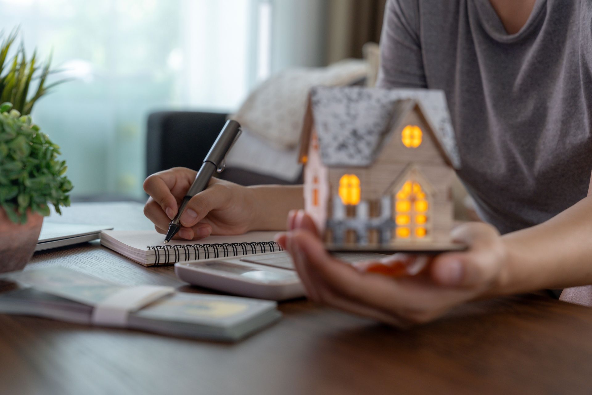 Person writing in notebook, holding miniature house, money and calculator on desk. Person writing in notebook, holding miniature house, money and calculator on desk.