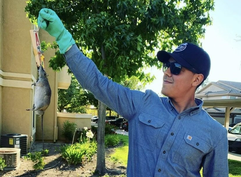 A man wearing a hat and sunglasses is holding a rat in his hand.