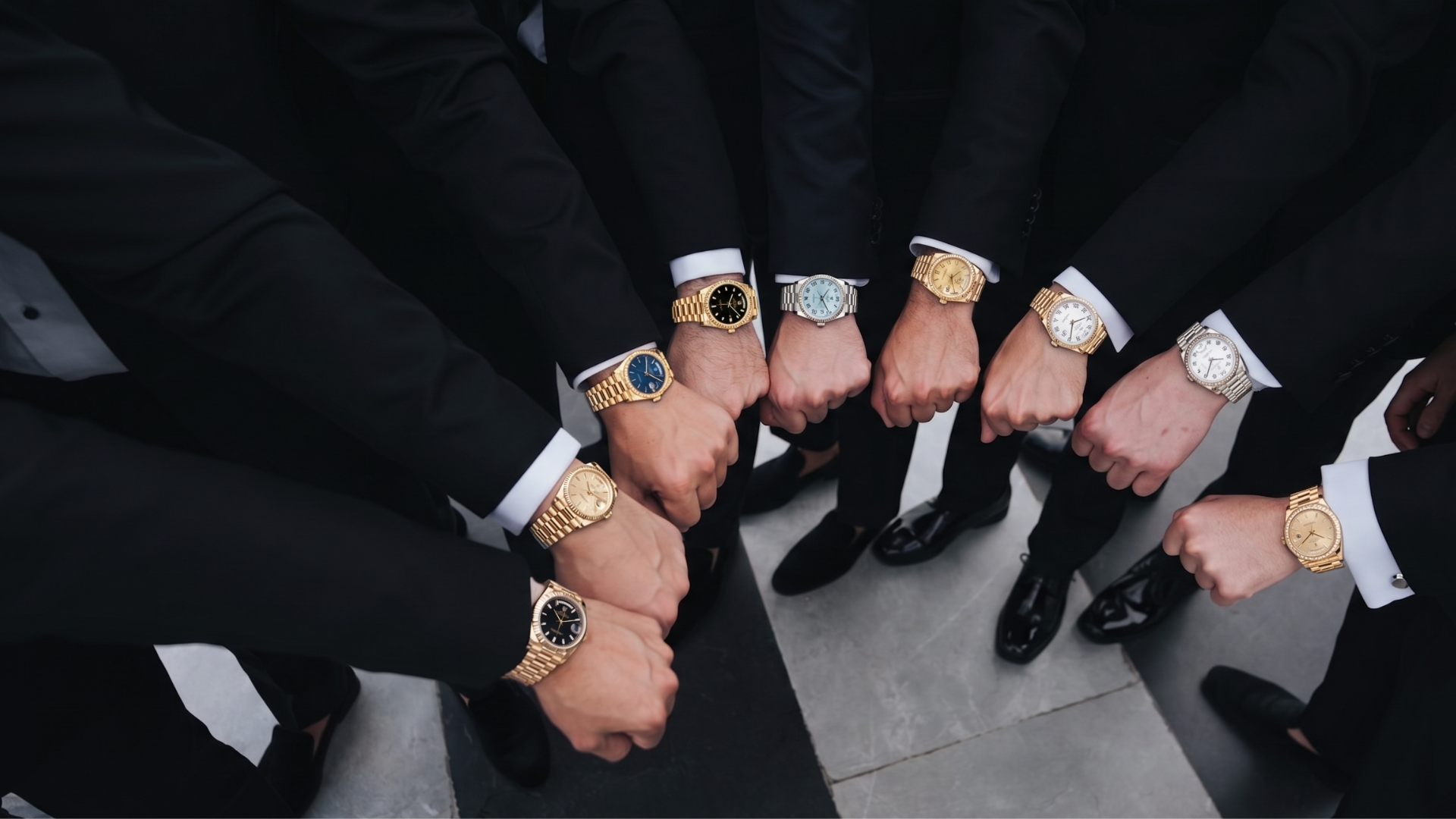Hands with luxury watches forming a circle over a tiled floor, all wearing black suits.