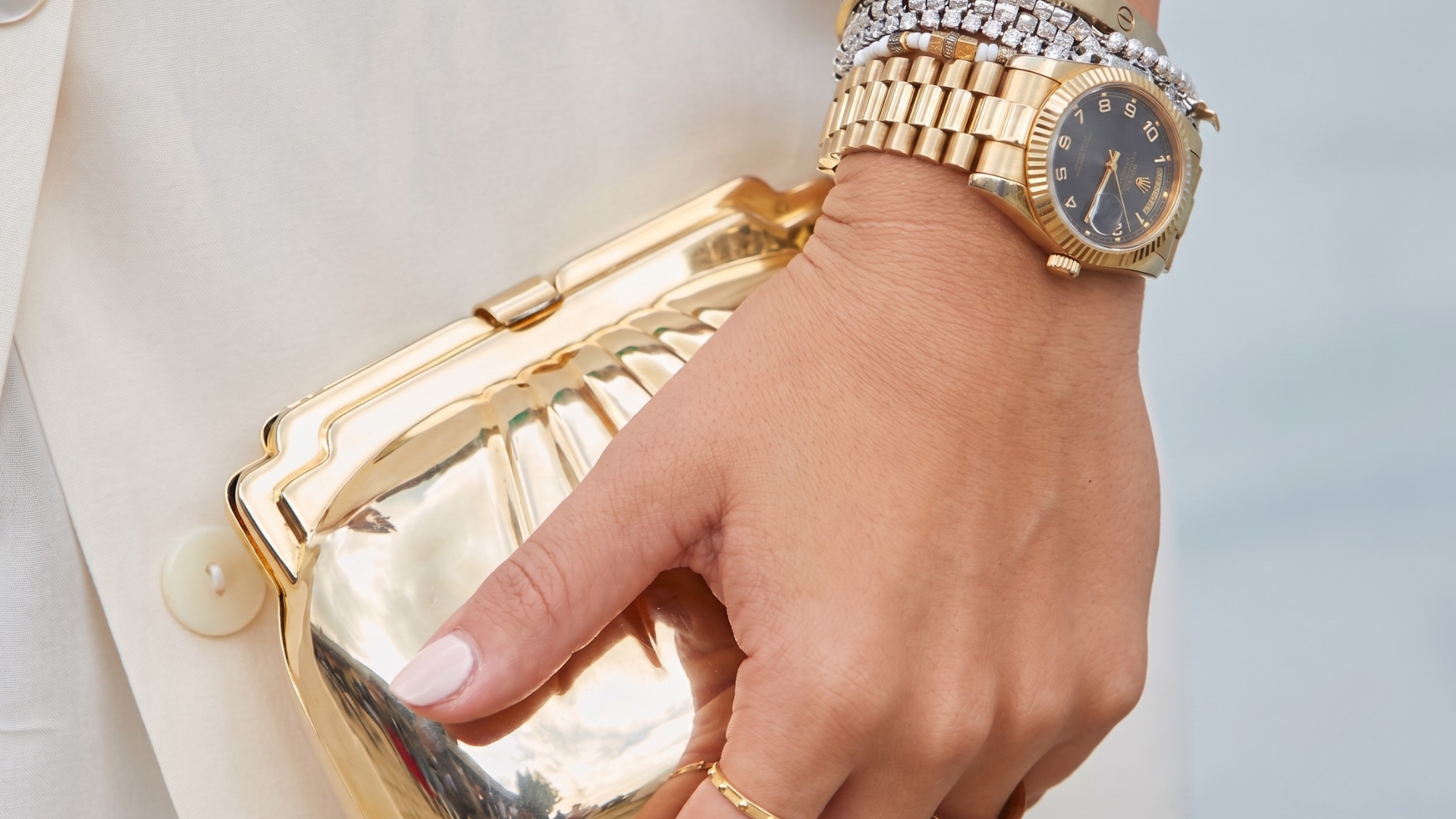 Hand holding a gold clutch beside a gold wristwatch and bracelet on a white outfit
