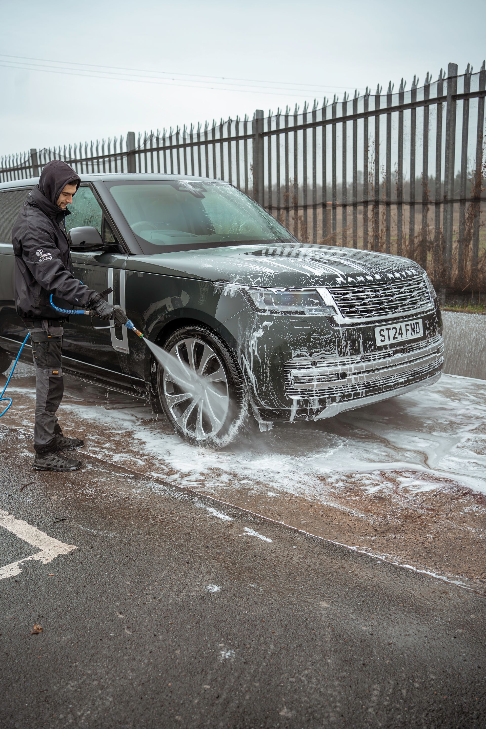 Person washing a black SUV with a pressure washer beside a metal fence on a wet roadside